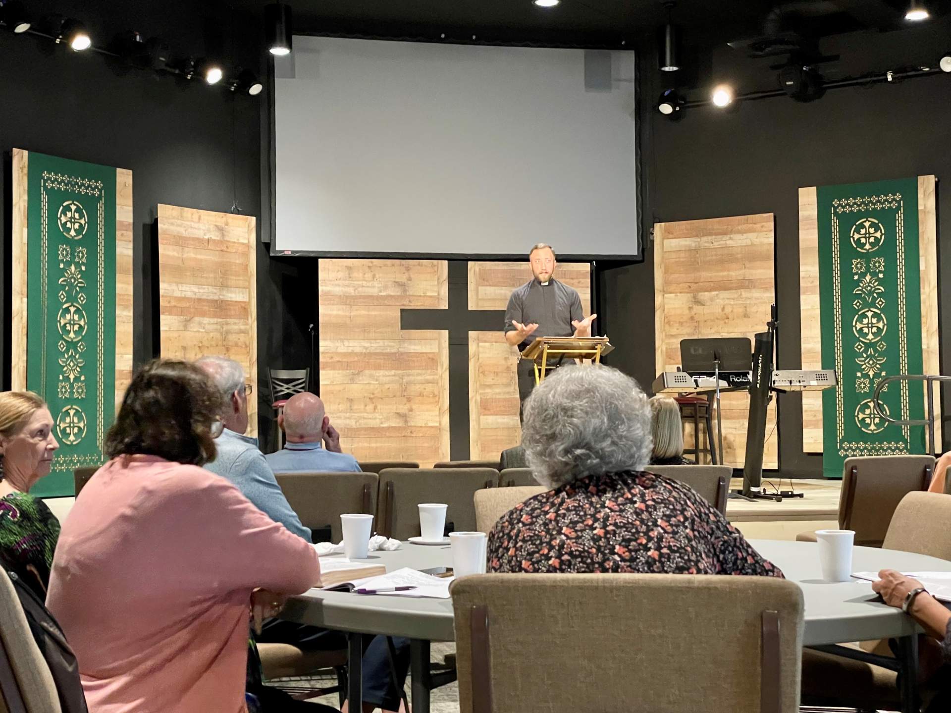 A man speaks at a podium in a church setting with a large cross behind him, as an audience listens.