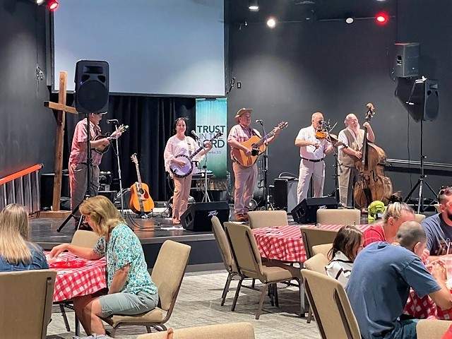 A folk band performs on stage in front of an audience seated at tables with red and white checkered tablecloths.